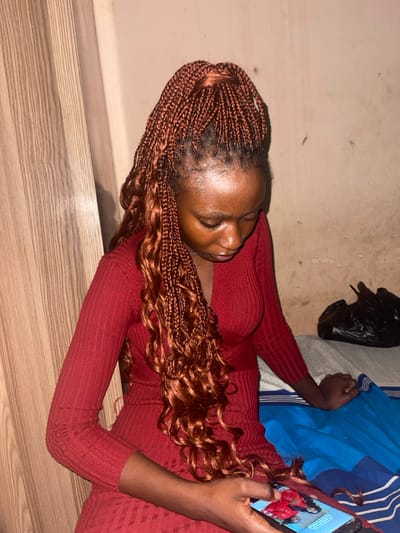 A woman in a red long-sleeve dress is lying down, possibly on the floor, next to a bed with blue and white striped sheets. Her head rests on the floor and she is holding a cell phone, looking at something on the screen. She has copper-colored braided hair with curly ends. A black plastic bag is visible on the bed behind her. The lighting is somewhat dim.