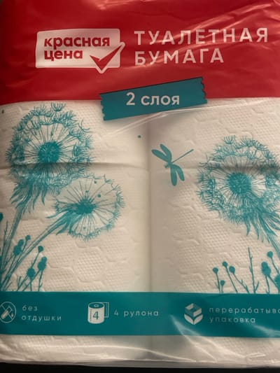 The image displays a package of toilet paper with a red label on the left and a teal blue label on the right, both vertically oriented. The toilet paper itself is white and embossed with a repeating pattern of dandelions and dragonflies in a teal blue color.

The red label features prominent white text in Russian. The largest text reads "ТУАЛЕТНАЯ БУМАГА" (TOILET PAPER). Below this, a white arrow-shaped emblem contains the text "красная цена" (red price).

The teal blue label has text running vertically. At the top, it reads "перерабатыва упаковка" (recyclable packaging). Below this is a symbol of a cube composed of smaller cubes. Further down, the text "4 рулона" (4 rolls) is present, accompanied by an icon of a toilet paper roll with the number 4 inside. At the bottom, "без отдушки" (unscented) is written, with a crossed-out fragrance symbol beneath it. There is also a smaller teal tab on the left side of the toilet paper with the word "слоя" (layer) printed on it, and a "2" next to it, likely indicating 2-ply.

The image appears to be taken indoors, likely in a retail setting or a home environment, given the product packaging. There are no people visible, and the focus is solely on the toilet paper product. The lighting is somewhat dim, suggesting it might not be natural daylight. The overall presentation is of a consumer product ready for sale or use.