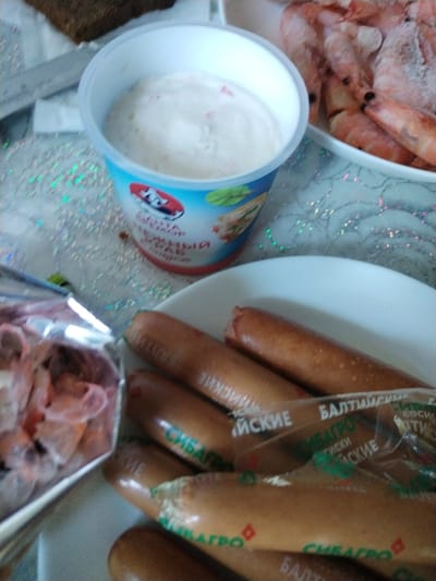 A close-up, overhead shot shows a food spread on a patterned tablecloth. In the foreground, a pile of cooked shrimp is partially visible in a silver container, while several sausages lie on a white plate. Some of the sausages are wrapped in clear plastic packaging with text in Russian. A white cup with a blue and red label containing a creamy white substance is positioned in the upper left quadrant. In the background, a piece of dark bread is visible. The lighting suggests it is daytime, and the setting appears to be indoors, possibly a home dining area.
The visible text on the sausage packaging reads "БАЛТИЙСКИЕ" and "СИБАГРО," which translates to "Baltic" and "Sibagro." The label on the cup also has Russian text, but it is largely illegible. The overall scene depicts a casual meal or preparation for one.