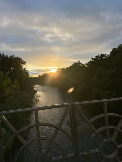 An outdoor, wide-angle, vertical shot captures a sunrise over a river, viewed from a bridge railing. The sun peeks through clouds in the upper center of the frame, casting golden rays across the scene. The river flows from the background to the foreground, its surface reflecting the sky and surrounding trees. Lush green trees line both banks of the river, creating a sense of depth. In the foreground, a metal bridge railing with a decorative, circular pattern is visible, adding a man-made element to the natural landscape. The overall impression is one of natural beauty at the start of a new day. The location is the River Severn in Broseley, United Kingdom.