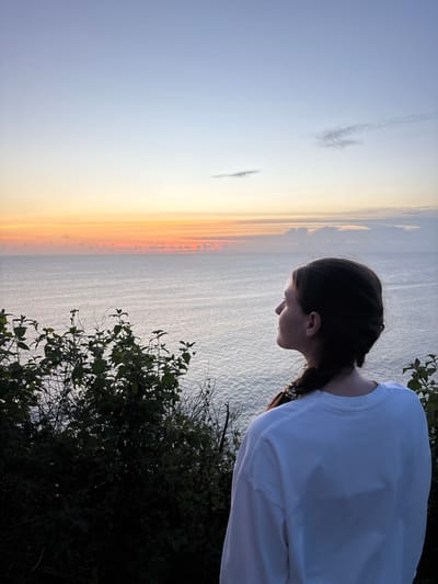 The image captures a person standing on a cliff overlooking the ocean at sunset. The individual, seen from the back and side profile, has brown hair styled in a braid. They are wearing a white, long-sleeved shirt. The background shows a coastal scene with the ocean reflecting the warm colors of the sunset. Vegetation grows along the edge of the cliff. The location is most likely Pecatu, Indonesia, based on the presence of the cliffside vegetation and ocean views.