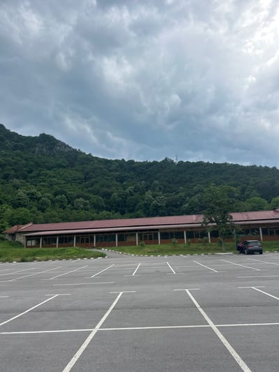 The image depicts an outdoor scene in Bachkovo, Bulgaria, featuring a large asphalt parking lot in the foreground, a long building, and a densely forested mountain under an overcast sky. The sky is filled with varying shades of grey clouds. A steep, verdant mountain covered in deciduous trees dominates the background, with a distinct rocky outcrop visible on its upper left slope and a power line structure on the ridge to the right.

A single-story building with a reddish-brown, possibly metal or tiled, roof is situated behind the parking lot. The building's facade consists of a section with visible stone or light-colored masonry on the far left, transitioning to a longer segment featuring numerous evenly spaced, large windows or glass doors with dark frames, supported by pillars. Green grass and low vegetation grow on a gentle incline between the building and the parking lot.

The parking lot is marked with white lines defining numerous "T" shaped parking spaces and appears mostly empty. On the far right side of the lot, a dark grey SUV, identifiable as a BMW X5, is parked. Its Bulgarian license plate, displaying "H 5645 MX," is visible. Two individuals are partially visible near the open rear passenger door of the SUV; one is wearing a pink garment, suggesting an interaction with the vehicle. A single, medium-sized tree with green foliage stands in a planted area within the parking lot, to the left of the parked SUV.