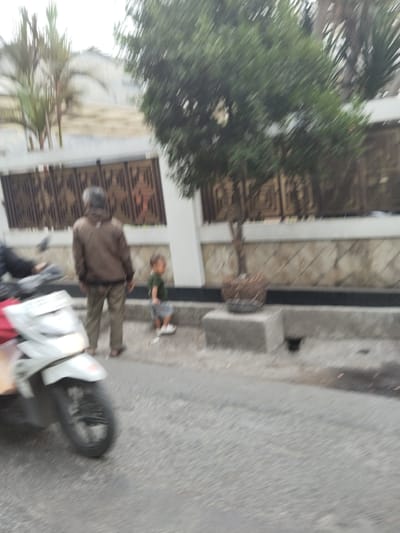 The image depicts an outdoor scene in what appears to be a residential area or along a street. In the foreground, a white motorcycle is visible on the left, partially blurred due to motion. Behind the motorcycle, a person wearing a brown jacket and khaki pants stands facing away from the camera. To the right of this person, a young child wearing a green shirt and a grey skirt or shorts is standing. The child is facing towards the right of the frame. In the background, there is a wall with a decorative pattern and some greenery, including a tree in a pot. The ground is paved with asphalt, and a concrete curb is present. The overall image is somewhat blurry, suggesting it was taken with a moving camera or in low light conditions. Based on the provided metadata, the location is identified as Bandung City, Indonesia.