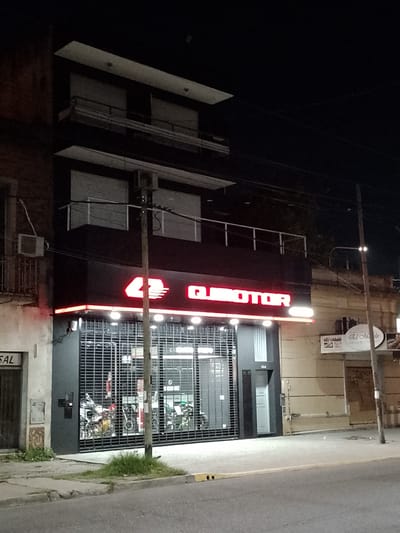 Nighttime view of a closed motorcycle shop, "Quimotor," located at number 484 in Quilmes, Argentina. The shopfront is secured with a black metal roll-down gate, partially revealing motorcycles inside. The building is three stories high, with the shop occupying the ground floor. The upper floors have closed roller shutters. A red neon sign illuminates "Quimotor" above the entrance. A wooden utility pole stands on the right side of the frame. Adjacent buildings include a restaurant named “El Almada”. The street is visible, with a small patch of vegetation growing near the curb.