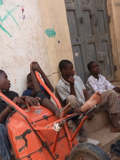 This image, taken in Kawaji, Nigeria, displays four young males in an outdoor urban setting. In the foreground, an orange metal wheelbarrow lies on its side, its single rubber wheel visible at the bottom right. A boy, wearing a blue shirt and dark trousers, holds the wheelbarrow's handles, with his bare feet resting on its inner frame. To his left, another boy in a dark shirt is partially visible, facing inward. To the right of the wheelbarrow, a third boy, dressed in a light-colored short-sleeved collared shirt, is seated against a beige wall marked with extensive green and other colored graffiti and stains; he appears to be holding a small dark object near his mouth. His bare feet are visible, resting on the ground near the wheelbarrow's framework. Further right, a fourth boy, wearing a white patterned long-sleeved shirt, is seated on concrete steps. The background features the aforementioned marked wall and a weathered, grey-blue double wooden door with visible metal hardware. A dark, possibly plastic, object is on the ground near the steps on the far right.