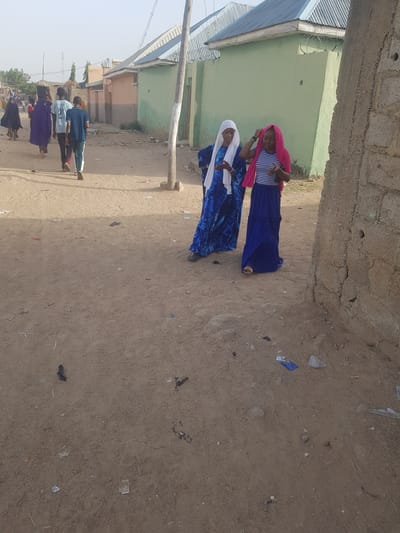 In Dabawa, Nigeria, two women walk along a dirt street enclosed by building walls. One wears a white headscarf and a blue patterned dress. The other wears a pink head covering, a striped shirt, and a blue skirt. Other people walk further down the street in the distance. Trash litters the dirt street. Buildings with corrugated metal roofs are visible.