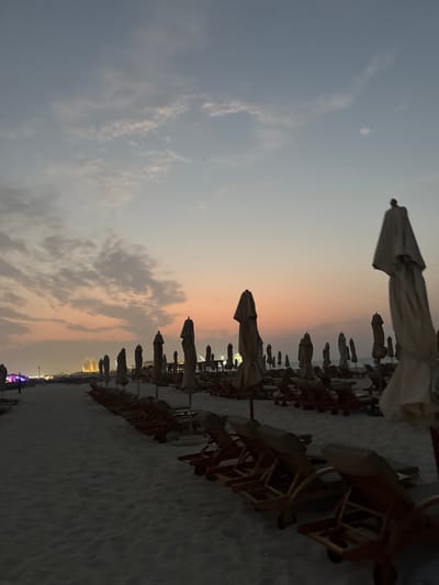 The image displays an unoccupied beach scene at dusk or sunset in Abu Dhabi, United Arab Emirates, with distant structures resembling the Etihad Towers visible on the left horizon, indicating a view towards the Corniche area. The foreground and midground are dominated by numerous rows of closed, light-colored beach umbrellas and associated empty wooden beach loungers, uniformly arranged on light-colored sand. A sandy path is visible on the left side of the frame. No individuals are present. The sky transitions from a soft orange and pink hue near the horizon to a lighter blue-grey overhead, featuring scattered, indistinct cloud formations. Faint city lights are visible along the distant left horizon.