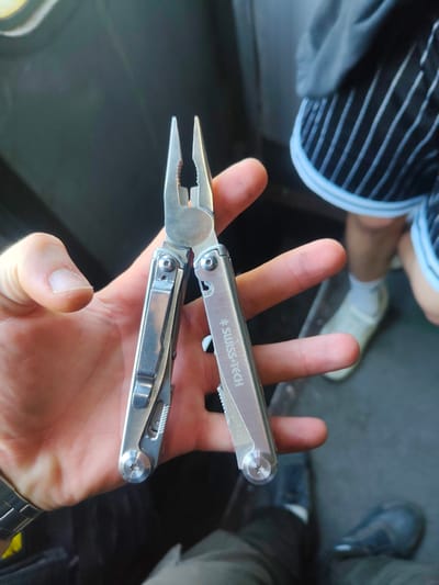 In Sviatopetrivske, Ukraine, a person's hand is holding a silver multi-tool, likely a Leatherman or similar brand, with the pliers extended. The multi-tool has the "SWISS+TECH" logo visible on its handle. The hand is positioned against a dark, blurry background, with hints of a person's legs and feet, wearing white socks and casual shoes, in the upper right portion of the frame. The overall lighting suggests an indoor or shaded outdoor environment.