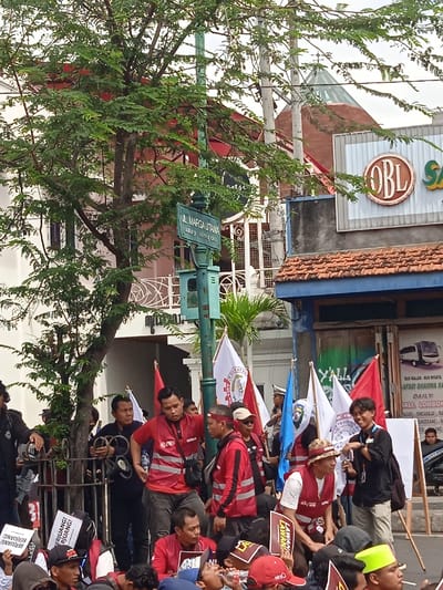 The image shows a street scene in Yogyakarta, Indonesia, at the intersection of Jl. Marga Utama and Jl. Affandi, as indicated by the street signs. The scene is populated by a crowd of people gathered around a street pole. The individuals are wearing red and white shirts with reflective vests, and many are holding flags and banners. There are also several people wearing black t-shirts and hats. The group is standing in front of a storefront with a blue awning and a sign with a red circle and the acronym OBL. The storefront is also adorned with a banner promoting a bus service, as well as a sign with the word “Yalla” on it. There is also a red-roofed building visible behind the crowd. The environment is urban and the scene is likely a rally or protest, with a focus on unity and potentially labor issues. The people are interacting with each other, holding up flags and signs, and listening to speakers, likely leaders of the group. The atmosphere appears to be energetic, enthusiastic, and potentially political, with the presence of a large crowd and various signs and banners. There is a large tree in the foreground, with a street pole in the center. The pole has a traffic light and a street sign with a small, green, and red circle. The scene appears to be captured on a sunny day, as the background includes a clear, blue sky.