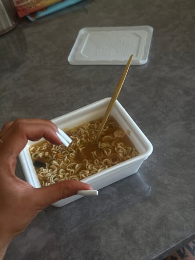 The image shows a top-down view of a hand holding a container of instant ramen noodles. The hand has long, white artificial nails. The ramen is in a white styrofoam container and appears to be broth-based with noodles and some green garnishes. A pair of wooden chopsticks is resting on the edge of the container, partially submerged in the broth. To the left of the ramen container, there is a white lid for the container, which has condensation on its surface. The scene is set on a textured, dark gray surface, possibly a table. The lighting suggests it is daytime. There is no visible text in the image. The location is identified as New Athos, Abkhazia.