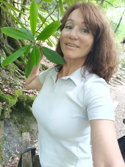 The image shows a woman with shoulder-length brown hair and blue eyes smiling at the camera. She is wearing a light blue, short-sleeved polo shirt. She is outdoors, holding a branch with green leaves in her left hand. In the background, there are trees, moss-covered rocks, and a dirt path. To the right, a blurred figure of a person in the distance is visible. The environment suggests a natural, wooded area, possibly a park or trail. The scene is set in Plastunka, Russia, a location known for its natural beauty and forests.