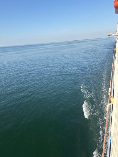 The image depicts a side view from a vessel at sea. The vast expanse of the ocean stretches out to the horizon under a clear blue sky. The water is a deep blue-green, with gentle ripples creating a textured surface. White foam trails indicate the wake of the vessel. On the right side of the image, the railing and side of the vessel are visible, featuring white and beige paneling and a thick rope. A life raft is mounted on the side of the ship. The scene is calm and expansive, with no discernible landmass or specific urban features. The location is identified as Unknown, Lithuania, suggesting the vessel is operating in Lithuanian waters.