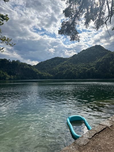 The image shows a tranquil lake scene with a backdrop of lush green mountains under a partly cloudy sky. In the foreground, a small, inflatable blue pool floats on the calm water near the rocky shore. The lake's surface is rippled, reflecting the sky and surrounding foliage. The scene evokes a sense of peace and natural beauty, characteristic of the Bavarian Alps, which surround Kiefersfelden, Germany. The location is likely a popular spot for recreation and enjoying the outdoors.