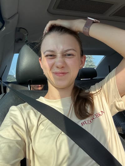 A woman is in the driver's seat of a car, her left hand is in her hair and her face is contorted in a playful grimace. She is wearing a light beige t-shirt with red text that reads "BULOCHKA." A man is visible in the backseat behind the headrest, his face partially obscured. The car's interior is visible, including the black leather seats and seatbelt. The background shows a landscape with trees and a blue sky, suggesting an outdoor location near Bürgstadt, Germany.
