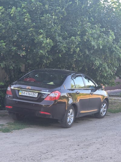 This media file captures a dark-colored car, likely a sedan, parked on the side of a gravel road. The car is the primary object, with a license plate that reads "BA 4164 BC" and the emblem "EMGRAND" visible on the rear. The car is positioned at an angle, with its rear towards the camera.

The scene is set outdoors, with lush green trees and foliage dominating the left side of the frame, suggesting a natural or semi-rural environment. The ground is a mix of gravel and sparse green grass. The lighting indicates it is daytime, with sunlight reflecting off the car's surface and filtering through the trees.

There are no people visible in the frame. The overall impression is of a static scene, with the car parked and no active event taking place. The location cues, such as the license plate and the mention of Sozonivka, Ukraine, place the image in a specific geographical context. The weather appears to be fair and sunny, suitable for outdoor activities. The image focuses on the vehicle and its immediate surroundings.