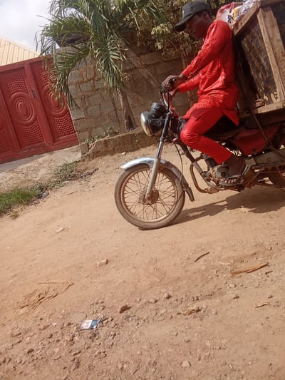 In Kagini, Nigeria, a person wearing a bright red patterned outfit and a black baseball cap rides a motorcycle. The motorcycle has a wooden cargo box attached to its rear, which is partially filled with various items. The person is seated on the motorcycle, gripping the handlebars, and their foot is resting on the footpeg. The ground is a dusty, unpaved surface with scattered rocks and vegetation. In the background, a red metal gate with intricate patterns and a brick wall are visible, along with a palm tree and foliage. The scene appears to be outdoors in a semi-urban or rural environment.