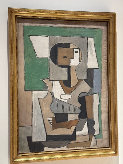 This is a full-frame photograph of a framed painting hanging on a white wall. The painting is in a Cubist style and features abstract geometric shapes and forms rendered in muted colors such as brown, grey, black, white, and a dull green. The composition appears to depict a figure or figures in a fragmented and deconstructed manner. The painting is housed in an ornate, gold-colored wooden frame with beaded detailing. The photograph is taken from a slightly elevated, straight-on angle, with the white wall and the top edge of the frame slightly out of focus in the background. The lighting is even and appears to be artificial gallery lighting, casting faint shadows on the wall behind the frame. There is no visible indication of the location within the city of Málaga, Spain.