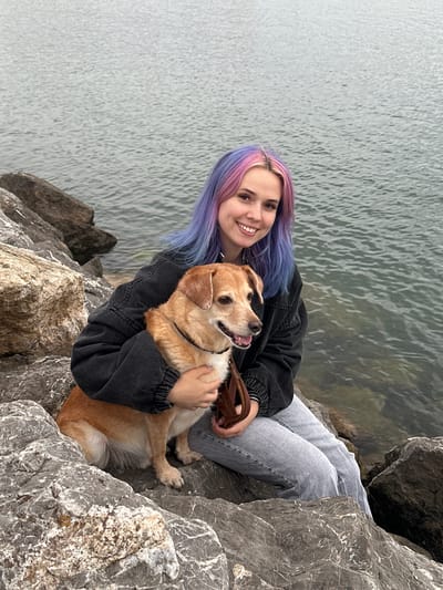 A young woman with split-dyed pink and blue-purple hair is seated on a rocky shore beside calm water in Gijón, Spain, holding a medium-sized tan and white dog. The woman wears a dark grey denim jacket and light-wash jeans, smiling directly at the camera with her arms wrapped around the dog. The dog, wearing a black collar and holding a brown leash, also looks towards the camera with its mouth slightly ajar. The foreground consists of large grey and brown rocks, extending to the edge of a flat, grey-green body of water.