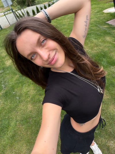 A young woman with brown hair and blue eyes smiles at the camera while outdoors. She is wearing a black crop top, black shorts, and white sneakers. A silver watch is on her left wrist and a silver necklace with two pendants hangs around her neck. She has a tattoo on her left arm that reads "this too shall pass" and another tattoo on her stomach. The woman is standing on a grassy area, likely a lawn, with a fence and trees visible in the background. The overall scene suggests a casual, outdoor setting. The specific location of Bistrica, Montenegro is not indicated within the image itself.