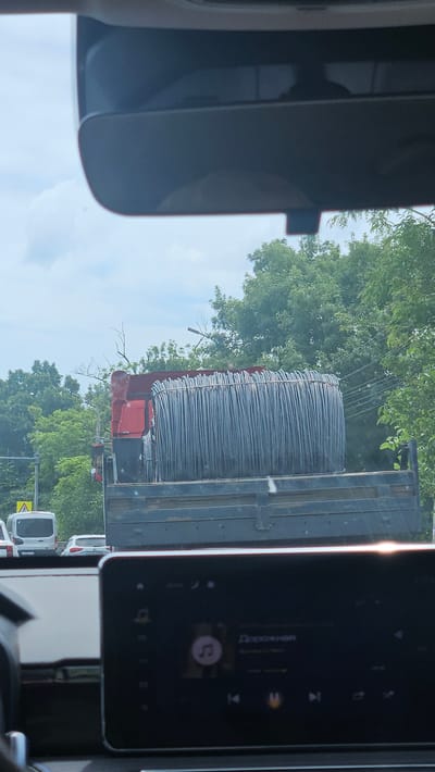 The image, taken from inside a vehicle, shows a large truck loaded with a massive coil of steel wire. The truck is red and black, and the coil of wire is silver and cylindrical, taking up most of the truck bed. The truck is on a road, with lush green trees and vegetation visible on either side of the road, suggesting an urban or suburban environment. The sky is overcast with some clouds. In the foreground, the dashboard of the vehicle is visible, with a dark screen displaying what appears to be music or media playback controls and text in Russian. A white van and another car are visible on the road behind the truck, indicating traffic. Based on the visible text on the car's display, the location is likely Novorossiysk, Russia.