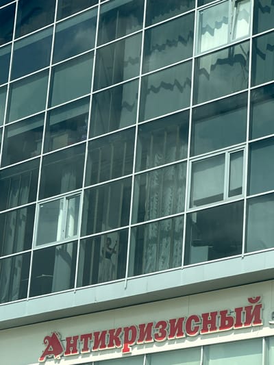 The image shows a close-up of a modern building with a glass facade. The building has a grid of white frames, dividing the glass into numerous windows. Reflections of clouds and wavy patterns are visible on the glass. In one of the windows, a person appears to be standing and looking at their phone. Below the glass facade, a sign with red Cyrillic lettering is visible. The sign reads "АНТИКРИЗИСНЫЙ" which translates to "Anticrisis". This building is located in Magnitogorsk, Russia.