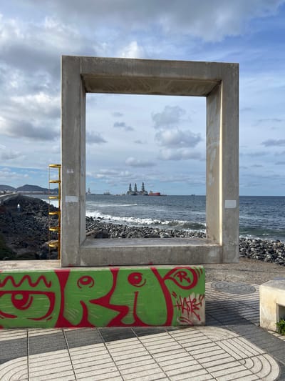 The image shows a view of the ocean framed by a concrete structure in Las Palmas, Spain. The ocean is visible through the square opening of the structure, with a rocky beach in the foreground. There are buildings and ships visible in the distance, further out in the ocean. To the right of the concrete frame, there's a painted or graffitied concrete support. The ground is a paved walkway with a gray and white geometric pattern. In the lower part of the view, below the ocean horizon, there is a rocky shoreline. The sky is overcast with clouds, with a portion visible above the concrete structure.