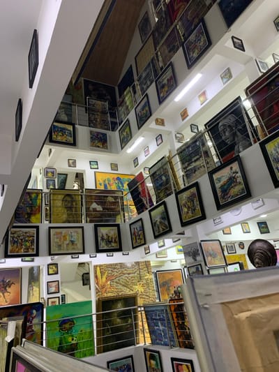 This image depicts a multi-level indoor art gallery located in Ikate, Nigeria. The environment is a brightly lit, white-walled space with numerous framed artworks displayed densely across all visible surfaces and levels. Metal railings delineate the different floors and staircases, creating an open, vertical layout. Fluorescent lighting fixtures are mounted on ceilings and under ledges, illuminating the extensive collection. A sloped, wooden ceiling section is visible towards the top.
The primary subjects are a vast quantity of paintings, varying significantly in size, style, and subject matter. Many works depict figurative scenes, including portraits of individuals, bustling market scenes, and landscapes. Abstract and semi-abstract compositions are also present. The frames are predominantly dark, likely black or brown wood. Paintings are hung on walls, displayed on shelves or ledges, and appear stacked against the metal railings on various levels. In the lower right foreground, a dark, sculpted bust with an elaborate headpiece is visible, positioned near other artworks. No live individuals are observed within the scene; the only human presence is through the depicted subjects in the artworks and the bust. The overall scene suggests a professional art exhibition or a commercial gallery space dedicated to showcasing a wide range of artwork.