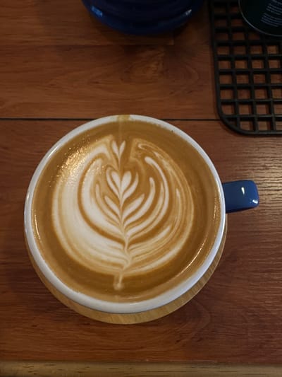 The image shows a close-up, overhead view of a cup of latte art on a wooden surface. The coffee has a creamy brown color with intricate white foam art resembling a fern leaf. A small, blue spoon handle is visible, sticking out of the top of the cup. To the left of the cup, part of a dark blue bowl or cup is visible. In the upper left corner, a portion of a black, grid-like object, possibly a placemat or coaster, is also present. The wooden surface appears to be a table with a visible grain. The lighting suggests an indoor setting, likely a cafe. No people are visible in the frame. There is no discernible time of day, weather, or specific location cues beyond the general setting. No text is visible in the image.