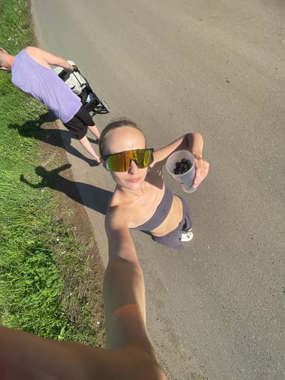 In the city of Podshivalovo, Russia, on a paved road bordered by lush green grass and wild flowers, a woman is captured in a selfie, holding a transparent cup filled with dark berries. She is wearing a grey sports bra and matching sweatpants, with distinctive white crocs on her feet. Her mirrored sunglasses reflect the bright sunlight. Behind her, a man in a lavender tank top and black shorts pushes a light-colored stroller along the road. The scene is illuminated by the sun, casting long shadows on the ground, indicating it is likely late afternoon or early morning. The overall impression is of a casual outdoor activity, possibly a walk or a stroll, with the berries suggesting a potential stop for foraging or a snack.