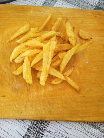 A pile of golden french fries is arranged on a wooden cutting board. The cutting board is positioned on a table covered with a black and white checkered cloth. The french fries are thin and appear to be freshly cooked, with some lighter and darker brown pieces scattered throughout the pile. The wooden cutting board has visible grain and some slight scuff marks. The image is taken from a top-down perspective.