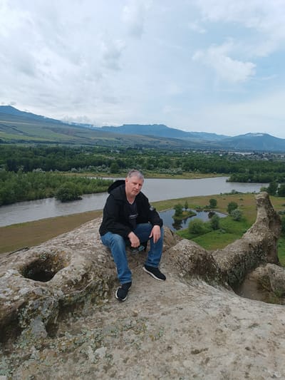 A middle-aged man with graying hair sits on a large, rough-textured rock with lichen growth, facing the camera. He wears a black jacket, a t-shirt, blue jeans, and black sneakers. His arms are resting on his knees, and he holds a pair of sunglasses in his right hand. The man is positioned in the foreground of the image, with his legs extending towards the right edge.
Behind him, a vast landscape unfolds. A wide, murky river flows from the left to the right, bordered by a grassy bank. Beyond the river, a dense forest of green trees stretches across the middle ground. In the distance, a town or city with buildings can be seen nestled amongst the trees, and further back, rolling hills and mountains under a cloudy sky complete the scene. The rock formation the man is sitting on appears to be part of a larger cliff or ancient structure, with a noticeable circular indentation or hole in the rock to his left. The environment suggests an outdoor, natural setting, likely a historical or scenic location in Uplistsikhe, Georgia.