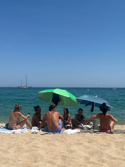 A group of six individuals is seated on a sandy beach in Badalona, Spain, near the Mediterranean Sea. Two umbrellas, one green and one blue, provide shade. The water is a vibrant turquoise, and several sailboats are visible in the distance. The sky is a clear, bright blue. The people are dressed in beach attire, with some appearing to be engaged in conversation or relaxing. The scene suggests a warm, sunny day at the beach.