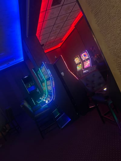 The image captures the interior of what appears to be a gaming or entertainment establishment, potentially located in Elena, Bulgaria, characterized by a combination of red and blue neon lighting. On one side of the frame, there is a series of illuminated arcade or slot machines with screens displaying colorful graphics. A button-tufted chair sits near the machines. The room has patterned walls and flooring. Red neon lights highlight architectural details. Blue neon lights illuminate the bar area. The image is presented at an angle, adding a sense of dynamism to the scene.