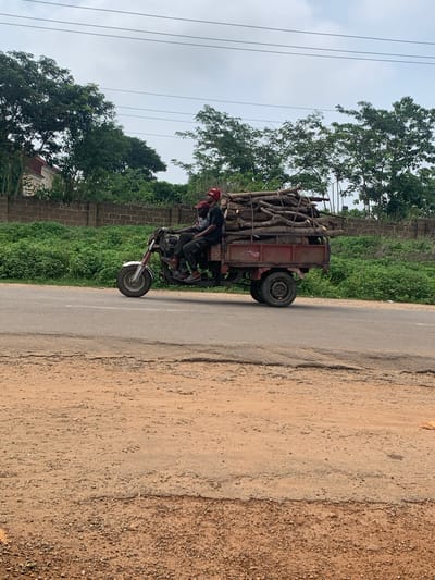 The image, taken in Gbeleko, Nigeria, captures a motorized tricycle heavily laden with cut wood or firewood. The vehicle is on a road that transitions from a paved surface to a dirt and gravel shoulder on the right.
Two individuals are on the motorcycle part of the tricycle. The person operating the vehicle wears a red cap, and another person is seated behind them. The cargo bed, a reddish-brown metal structure, is packed with logs and branches of varying sizes, piled high, indicating a significant load. The motorcycle features a silver front wheel fender.
The scene is set outdoors during the daytime under an overcast or cloudy sky. Lush green trees and dense vegetation form the background, and a brick wall is visible along the roadside. Several power lines stretch across the sky in the upper portion of the image. There is no visible text identifiable on the vehicle or within the environment. The image portrays a common sight of local transportation and resource gathering in the region.
