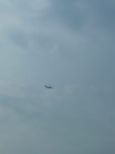 An airplane is silhouetted against a hazy sky. The aircraft is seen from its side, with its wings extended and tail clearly visible. It appears to be in mid-flight. The background is an overcast sky with diffused light, suggesting it might be dawn, dusk, or a cloudy day. The location is in the city of Kosambi Barat, Indonesia.