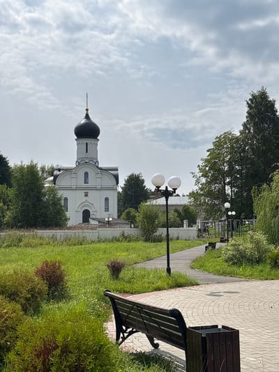 The image shows a white church with a black dome and a cross on top, set against a cloudy sky. In the foreground, there is a paved walkway with a park bench and a trash can. Lush green grass and trees surround the church and the walkway. The scene appears to be in a park or a serene outdoor area in Breytovo, Russia.