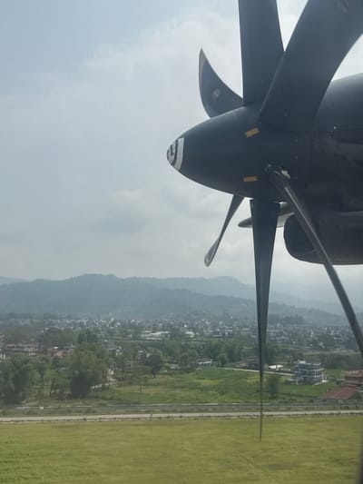 The image, taken from an aircraft, features a turboprop engine with its propeller blades prominently in the foreground. The aircraft is in flight over the city of Pokhara, Nepal. The landscape is a mix of urban development and green spaces. A runway is seen to the right of the frame, adjacent to a road. Beyond the city, a range of mountains is visible in the distance. The sky is overcast, suggesting hazy or cloudy conditions.