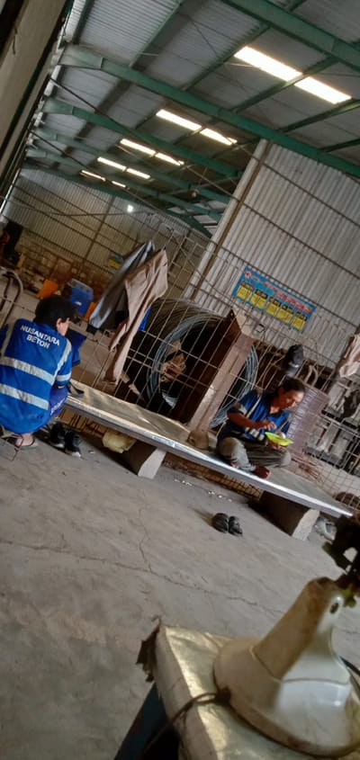 The photograph shows the interior of a metal warehouse, possibly in Sumput, Indonesia. Two men are present. One man wears a blue work jacket with the words "NUSANTARA BETON" printed on the back. He crouches near the edge of a raised metal platform supported by concrete blocks. The other man sits cross-legged on the same platform, eating food from a green plate. Pairs of sandals and shoes are discarded on the floor nearby. The warehouse structure is made of corrugated metal, supported by green metal beams. A large wire cage filled with coiled metal wire is visible behind the platform. There are additional objects scattered around, including jackets hanging and blue containers. A dirty, white object sits on what appears to be a bag, in the foreground. The floor is concrete. The lighting is bright, presumably from skylights in the roof.
