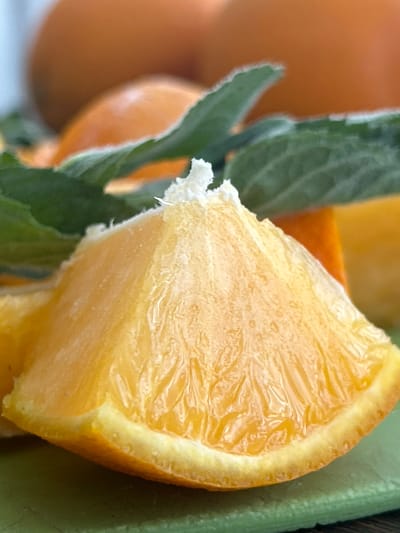 A close-up, eye-level, vertical shot focuses on a bright orange segment that fills the foreground, resting on a green textured surface, likely a cutting board. The orange segment is detailed, revealing its juicy pulp and fibrous texture. Behind it, out of focus, are more oranges and green mint leaves. The lighting is soft, creating a slight sheen on the fruit. The image does not depict any elements that would suggest a location in Kemerovo, Russia.