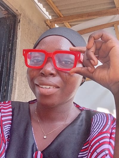 The scene is situated in the city of Ayob, Nigeria. It features a close-up portrait of an adult female with dark skin, positioned frontally and smiling, revealing her upper teeth. She wears a black head covering and a garment with a black V-neck bodice. The sleeves and shoulders of the garment are patterned with red, purple, and white wavy stripes. A thin gold-colored chain with a small, clear, spherical pendant is visible around her neck. Her left hand is raised, holding a pair of red, thick-framed glasses with clear lenses in front of her eyes. The background includes a light-colored, textured wall, a dark doorway opening to the left, and overhead exposed wooden structural beams supporting corrugated metal sheeting. The area is illuminated by natural light.