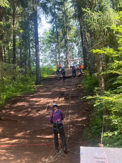 The image depicts an outdoor adventure activity, likely a ropes course or zip-lining event, taking place in a wooded area. Several individuals, dressed in athletic attire and safety harnesses, are engaged in ascending or preparing to ascend a natural slope or climbing structure.
The primary subjects are the people participating in the activity. One person in the foreground wears a purple jacket and black pants, with a blue helmet. Another individual further up the slope wears an orange and blue outfit. Multiple other figures in similar gear are visible, suggesting a group activity. Objects of note include ropes, carabiners, and safety harnesses, essential equipment for such activities. The setting is a forest with tall trees and dense foliage, with a dirt path or slope forming the ground.
The activity appears to be a form of climbing or ascending challenge. The presence of ropes and harnesses indicates safety measures are in place. The lighting suggests it is daytime, likely with clear weather given the bright sunlight filtering through the trees and casting shadows.
A sign is partially visible on the right side of the image, featuring white material with red text. The visible text appears to be in Cyrillic script and reads "Подъём по склону," which translates to "Climbing the slope." This text confirms the nature of the activity. The overall mood appears to be one of focused effort and potentially excitement, typical of outdoor adventure sports. The location context of Сергино, Russia, provides a geographical reference for the scene.