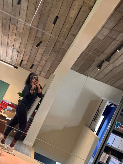 In Maldegem, Belgium, a forensic analysis of the provided image reveals an indoor mirror selfie. A woman with dark hair in a high ponytail, dressed in a black long-sleeved top, black leggings, and light-colored sneakers with brown socks, stands before a large, multi-paneled mirror, capturing her reflection with a smartphone held to her face, obscuring her features. She wears a watch and a gold-colored necklace.
The reflection in the mirror displays a rustic wooden plank ceiling with visible grain, adorned with multiple black track lights, several of which emit a warm glow. Thin wires or tension cables are strung horizontally across the ceiling. Below the ceiling, light-colored (beige/off-white) walls or architectural elements are visible, including a prominent diagonal beam or panel reflecting the wooden ceiling.
In the background of the reflection, to the left of the woman, a dark green rectangular panel is mounted on a light wall. Beneath this, several cardboard boxes and a red plastic storage bin are stacked on what appears to be a light-colored floor. A green potted plant is partially visible behind the woman. To her right, a tall, narrow white vertical structure stands. Further right, a doorway with a bright blue interior frame leads to another area, possibly revealing a window or light source within. Adjacent to the doorway, dark shelving units are stocked with neatly piled papers or books on lower shelves and small, colorful objects on the top shelf. The entire scene is captured with a slight upward and rightward tilt, indicating the angle at which the mirror selfie was taken.