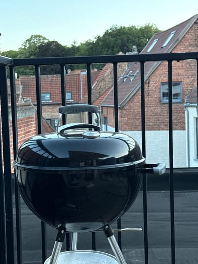 A black kettle grill is positioned on a balcony overlooking a cityscape of Bruges, Belgium. The grill is in the foreground, with its reflective surface showing an inverted image of the balcony railings and the sky. The background features traditional brick buildings with tiled roofs, characteristic of Bruges architecture. Dormer windows and a few scattered trees are visible. The scene appears to be set during the daytime, with a clear sky.
