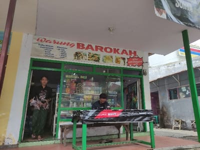 This image, rotated 90 degrees clockwise, shows a small Indonesian shop or "warung" from an elevated perspective, likely in Situbondo, Indonesia. The shop features a large display window framed in green, revealing shelves stocked with various packaged goods, possibly snacks and drinks, behind a counter.

Two individuals are visible through the shop window. A man is clearly reflected in the window, facing the viewer, while a woman wearing a patterned top and a green skirt stands further inside the shop. Both appear to be present for business, perhaps as owner or staff.

Below the window, a white banner prominently displays the shop's name, "Warung BAROKAH," in red letters. The banner also lists a menu of Indonesian dishes and beverages, including "Sedia Makanan" (Sells Food), "Nasi Rawon," "Pecel," "Kare," "Campur," "Lalapan," "Es Teh," "Jeruk," "Josua," and "Kopi." A smaller sign above the window also reads "WARUNG NASI" (Rice Stall). Additional text, "SULTRA PERLENGKAPAN BATI," suggests the sale of batik-related items or a broader selection of goods.

The surrounding area consists of simple, concrete buildings, with a street or alleyway visible in the background, reinforcing the local, urban setting. A portion of a blue and red sign is visible in the lower left corner of the background. The scene depicts a typical neighborhood shop, likely open for daily business.