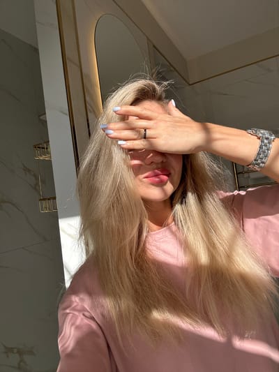 An outdoor medium shot shows a woman with blonde hair covering her eyes with her hand. She is wearing a pink shirt and a watch on her left wrist. A ring is visible on her left ring finger. The woman's lips are slightly parted. She is standing in front of a wall with white and gray marble patterns. A shelf unit is attached to the wall on the left side of the image. Sunlight is casting shadows on the woman and the wall. The background is out of focus, but appears to be a bathroom or a room with similar decor. The scene is not identifiable as being in Armavir, Russia.