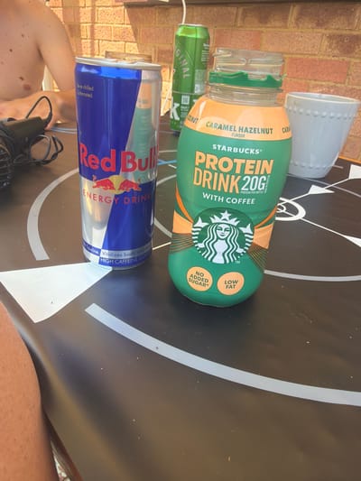 The image shows a close-up of a Red Bull energy drink can and a Starbucks caramel hazelnut protein drink bottle on a black and white patterned surface, possibly a table. A white ceramic pot is placed near the bottle. Part of a brick wall is visible in the background. A microphone and cable are visible near the Red Bull. Part of a human body, including skin on the shoulder and leg are included in the frame. The location is unknown and cannot be determined as Swindon, United Kingdom, from the provided images.