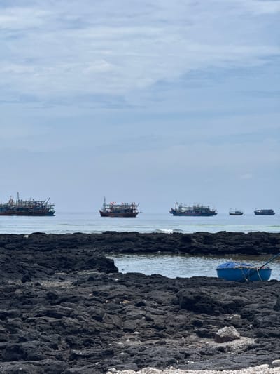 The image captures a view of the sea near Lý Sơn Island, Vietnam, characterized by a rocky foreground and a calm sea dotted with several fishing boats. The foreground is dominated by dark, uneven volcanic rocks, indicative of Lý Sơn's volcanic origin. A small blue boat, possibly a buoy or a small dinghy, rests on the rocks near the water's edge. In the middle ground, several fishing boats of varying sizes are visible on the water, some appearing to be anchored or moving slowly. The sea is a light blue, merging with the hazy sky. The sky itself is overcast with light gray clouds, suggesting a temperate or cloudy day. The overall impression is one of a coastal fishing village or an area where maritime activities are prominent.