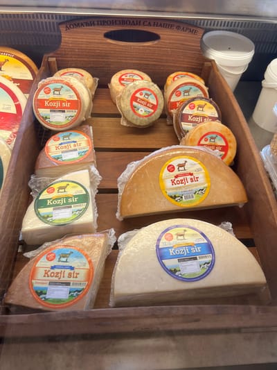 The image shows a close-up view of a display of goat cheese at a farm shop, likely in Kovačko Polje, Montenegro, based on the text. Several wheels and wedges of goat cheese are presented in wooden crates, with clear plastic packaging and labels indicating "Kozji sir" (goat cheese) and "Farma Carevic". The labels also feature graphics of goats and some nutritional information. In the background, white buckets are visible, and a wooden sign with Cyrillic script reading "ДОМАЋИ ПРОИЗВОДИ СА НАШЕ ФАРМЕ" (Homemade products from our farm) is positioned above the cheese display. The lighting suggests an indoor setting, possibly a refrigerated display case.