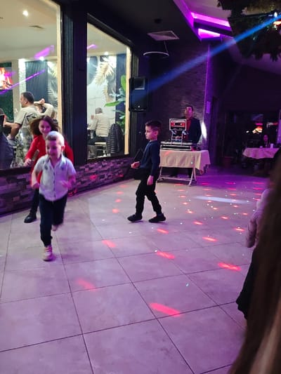 An indoor scene in Opaka, Bulgaria, depicts an event hall or restaurant under dim, ambient purple lighting with projected red and white disco light patterns on a light-tiled floor.

In the foreground, a young boy with light hair, wearing a light-colored collared shirt and dark trousers, is captured in motion, running towards the viewer's left. Behind him, slightly to the left, a girl with dark hair, dressed in a red top or dress, is partially visible. To the right of the running boy, another young boy, dressed in a dark blue long-sleeved shirt and black trousers, walks towards the right, looking slightly to his left.

In the mid-ground right, an adult male, likely a DJ, stands behind a white-skirted table equipped with professional audio mixing equipment and a red-faced stereo unit. He wears a dark jacket over a patterned shirt and observes the scene. A large, black speaker is mounted on the wall to his left. Overhead, a blue light beam cuts across the scene from the upper right.

The left side of the image features a large reflective surface, possibly a window or mirror, revealing an adjacent section of the venue. Reflections show several individuals, including a man in a light-colored shirt standing, and an older man seated at a table against a wall decorated with a tropical leaf pattern wallpaper. The ceiling is dark, featuring an air vent and a circular ring light fixture. Decorative foliage is faintly visible in the upper right background. Further in the background, to the far right, a dimly lit table with seated patrons is visible. A blurred portion of an individual's arm and shoulder is present in the extreme foreground right.
