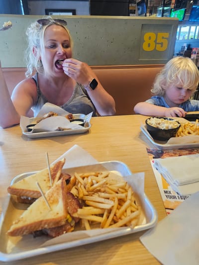 A photograph captures a woman and a young child at a restaurant in Homestead, Florida, United States. The woman, fair-skinned with blonde hair and sunglasses resting on her head, holds food in her hand, poised to eat it. A smartwatch adorns her left wrist, and she is wearing a gray tank top. Across the table, a blonde-haired child is visible, engaged with food.

Several serving trays of food are arrayed on the table. One tray contains French fries and pieces of toast secured with toothpicks, and another holds chicken wings with dipping sauce. A bowl of macaroni and cheese is also on the table. A wall with the number "85" in yellow is partially visible. The setting is indoors, lit by overhead lighting.